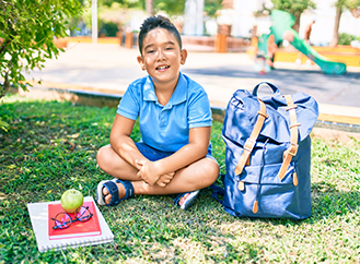 Happy elementary student sitting on grass next to his backpack